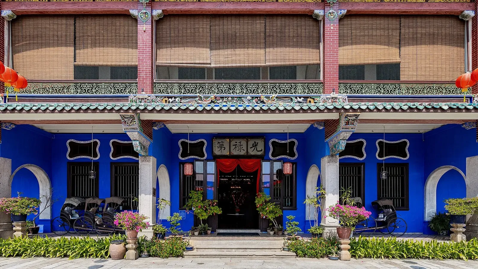 ด้านหน้า Blue Mansion ปีนัง หรือคฤหาสน์ Cheong Fatt Tze ผนังสีคราม ระเบียงชั้นสองมีราวเหล็กหล่อ และไม้กระถางเรียงหน้าบ้าน ภาพถ่ายเดือนมกราคม 2020