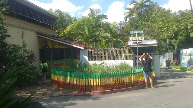 ลานใน Bob Marley Museum มีแปลงต้นไม้ล้อมรั้วไม้ระแนงทาสีแดง เหลือง เขียว ต้นปาล์มสูง และป้ายชื่อ Bob Marley Place ข้างอาคารพิพิธภัณฑ์ ภาพถ่ายเมื่อ 23 พฤศจิกายน 2019