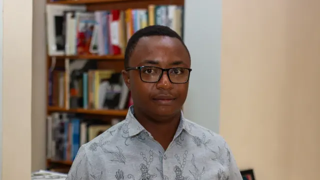 Simbarashe Steyn Kundizeza ผู้ชนะ Caine Prize 2026 สวมแว่นและเสื้อเชิ้ตลายพิมพ์ ยืนหน้าชั้นหนังสือ