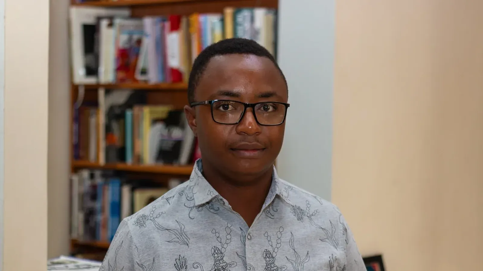 Simbarashe Steyn Kundizeza ผู้ชนะ Caine Prize 2026 สวมแว่นและเสื้อเชิ้ตลายพิมพ์ ยืนหน้าชั้นหนังสือ