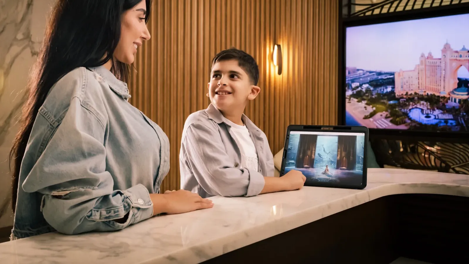 ภาพประกอบประกาศ Emirates Mile-A-Minute 2026 ของ Emirates แสดงแขกที่เคาน์เตอร์ต้อนรับ มีภาพโรงแรม Atlantis The Palm ในดูไบบนจอด้านหลัง