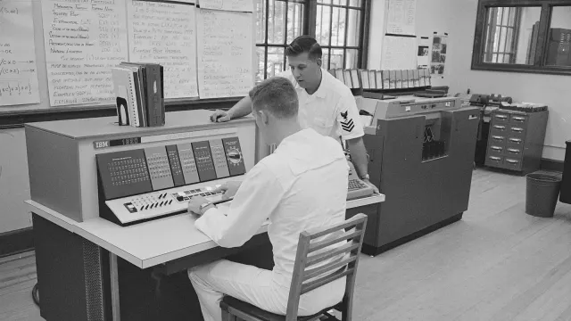 นักเรียนนายเรือนั่งใช้คอนโซลของ IBM 1620 โดยมีครูฝึกยืนดู ที่ U.S. Naval Academy เมื่อ 18 กันยายน 1963 ผนังด้านหลังติดแผ่นสรุปคำสั่งภาษา FORTRAN