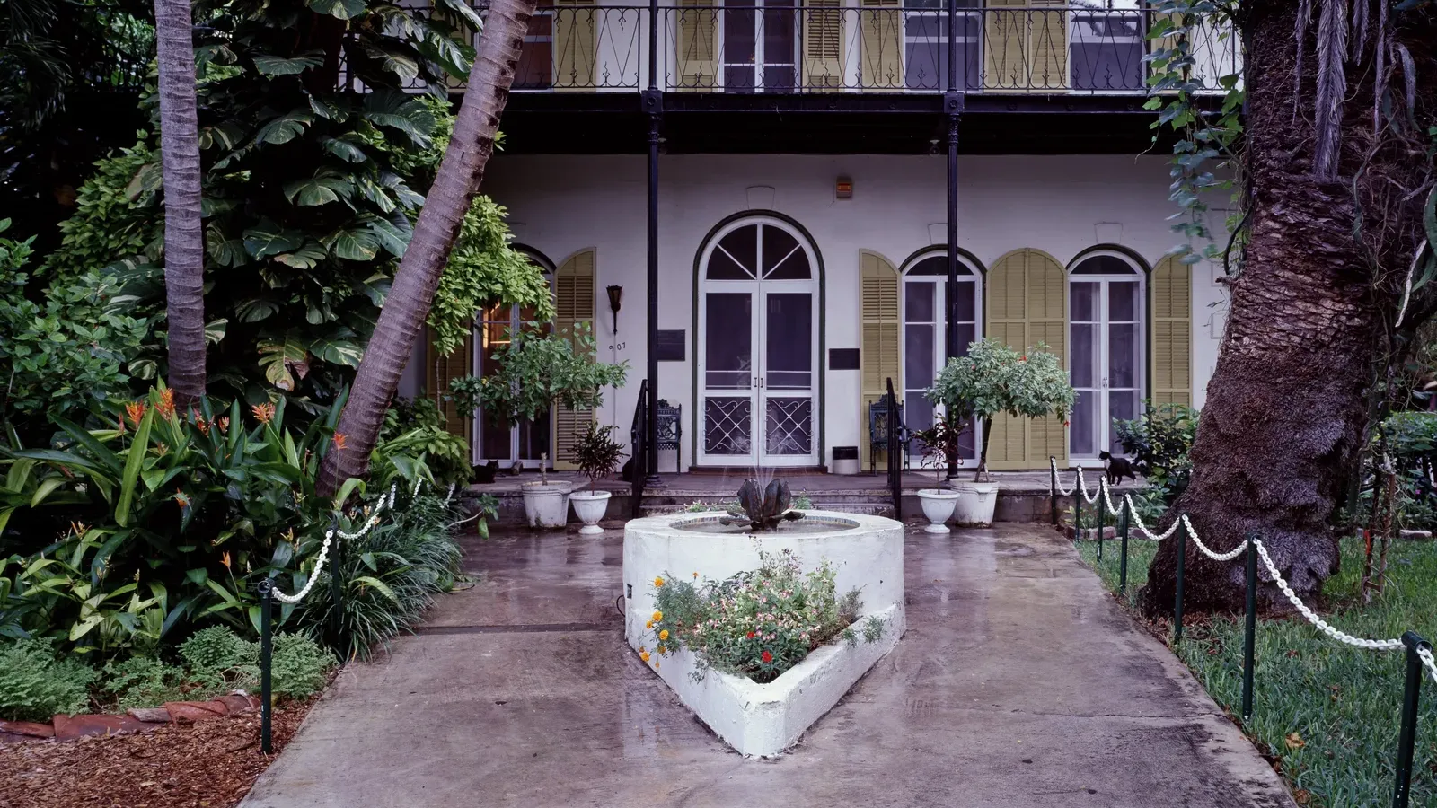 ทางเข้าหน้าบ้านเฮมิงเวย์ที่ Key West บ้านสองชั้นสีขาว บานเกล็ดสีเหลือง ระเบียงเหล็กชั้นบน และสวนไม้เขตร้อนสองข้างทางเดิน