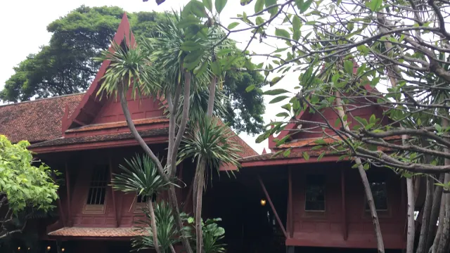 เรือนไม้สักทาสีแดงของบ้านจิม ทอมป์สัน หลังคาจั่วสูงแบบเรือนไทยสองหลังเชื่อมกัน มีรูปสลักหินคู่หน้าประตูและไม้ใหญ่ในสวนด้านหน้า