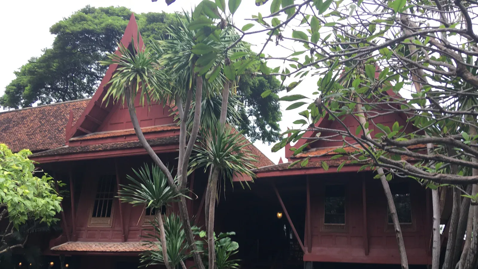 เรือนไม้สักทาสีแดงของบ้านจิม ทอมป์สัน หลังคาจั่วสูงแบบเรือนไทยสองหลังเชื่อมกัน มีรูปสลักหินคู่หน้าประตูและไม้ใหญ่ในสวนด้านหน้า