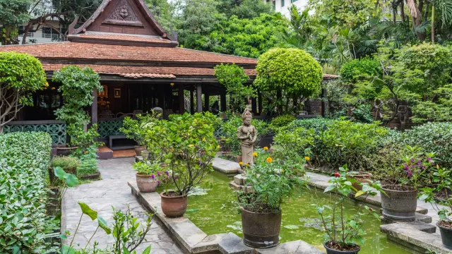 บ้านคึกฤทธิ์ ซอยสวนพลู เรือนไทยไม้หลังคาจั่วสูงมีหน้าจั่วแกะสลัก ด้านหน้าเป็นสวนที่มีสระน้ำ ไม้กระถาง และรูปปั้นหิน มีอาคารสูงอยู่ด้านหลัง