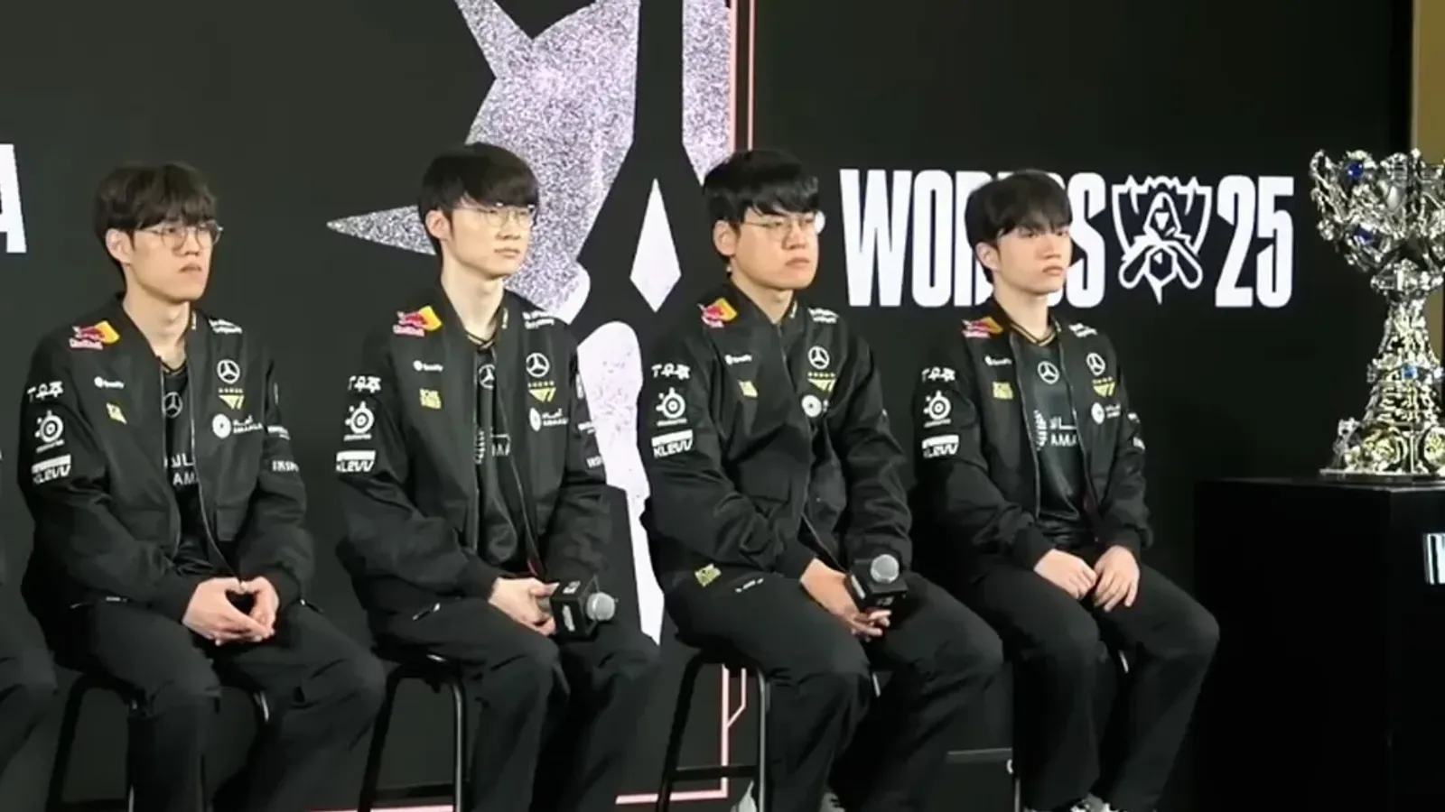 ผู้เล่นทีม T1 แชมป์เก่าในเสื้อแข่งสีดำนั่งบนเวทีงาน Media Day ของ Worlds 2025 ข้างถ้วยรางวัลแชมป์โลก ก่อนป้องกันแชมป์ใน LoL Worlds 2026 เวลาไทย