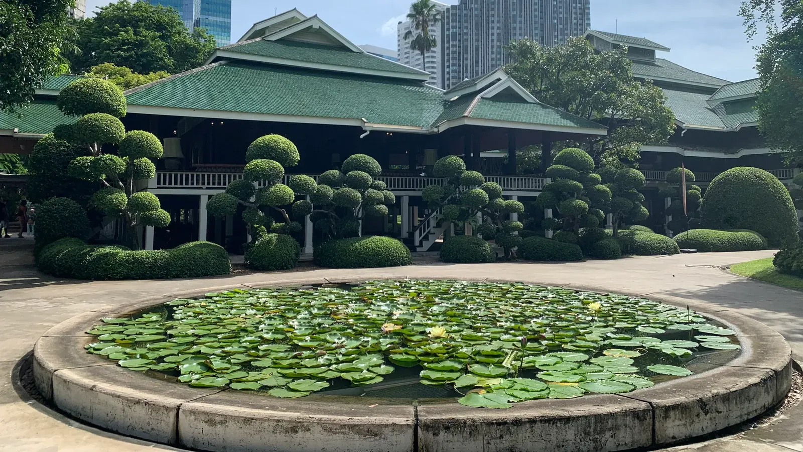 บ้านปาร์คนายเลิศ เรือนไม้หลังคาสีเขียวหลายชั้น มองจากอ่างบัวทรงกลมหน้าบ้าน มีไม้พุ่มตัดแต่งเป็นก้อนและตึกสูงย่านเพลินจิตอยู่ด้านหลัง