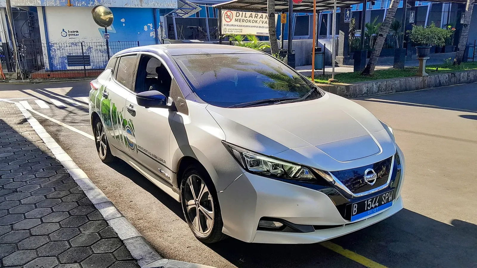 Nissan LEAF รุ่นที่ 2 (ZE1) สีขาวติดสติกเกอร์มหาวิทยาลัยบูดีลูฮูร์ จอดริมถนนภายในมหาวิทยาลัย มีนาคม 2024
