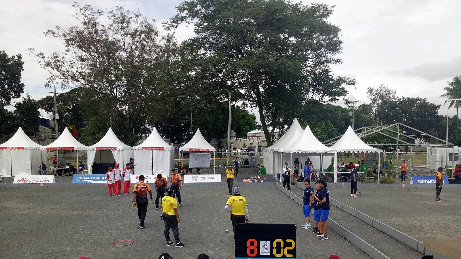ลานเปตองในซีเกมส์ 2019 ที่ Clark Freeport ฟิลิปปินส์ นักเปตองหญิงไทยในเสื้อโปโลสีเหลืองกางเกงดำแข่งประเภททีม 3 คนกับลาวในเสื้อสีกรมท่า มีวงโยนสีแดงบนพื้นกรวดและป้ายคะแนน 8-02