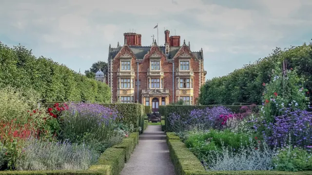 Sandringham House ตัวบ้านอิฐแดงมองจากทางเดินในสวน ขนาบด้วยแปลงไม้ดอกขอบต้นบ็อกซ์และแนวต้นไม้ตัดแต่งสูง