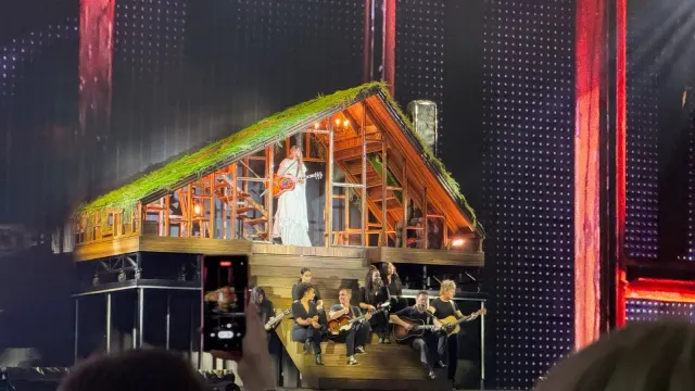 Taylor Swift ยืนถือกีตาร์ในฉากบ้านไม้หลังคามอสบนเวที The Eras Tour ช่วง folklore ที่เมือง Gelsenkirchen ประเทศเยอรมนี วันที่ 17 กรกฎาคม 2024 นักดนตรีนั่งเล่นกีตาร์บนบันไดหน้าฉากบ้าน