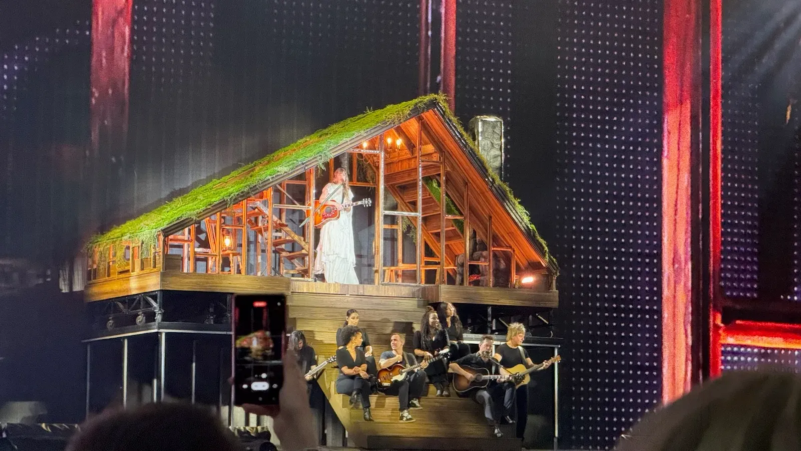 Taylor Swift ยืนถือกีตาร์ในฉากบ้านไม้หลังคามอสบนเวที The Eras Tour ช่วง folklore ที่เมือง Gelsenkirchen ประเทศเยอรมนี วันที่ 17 กรกฎาคม 2024 นักดนตรีนั่งเล่นกีตาร์บนบันไดหน้าฉากบ้าน