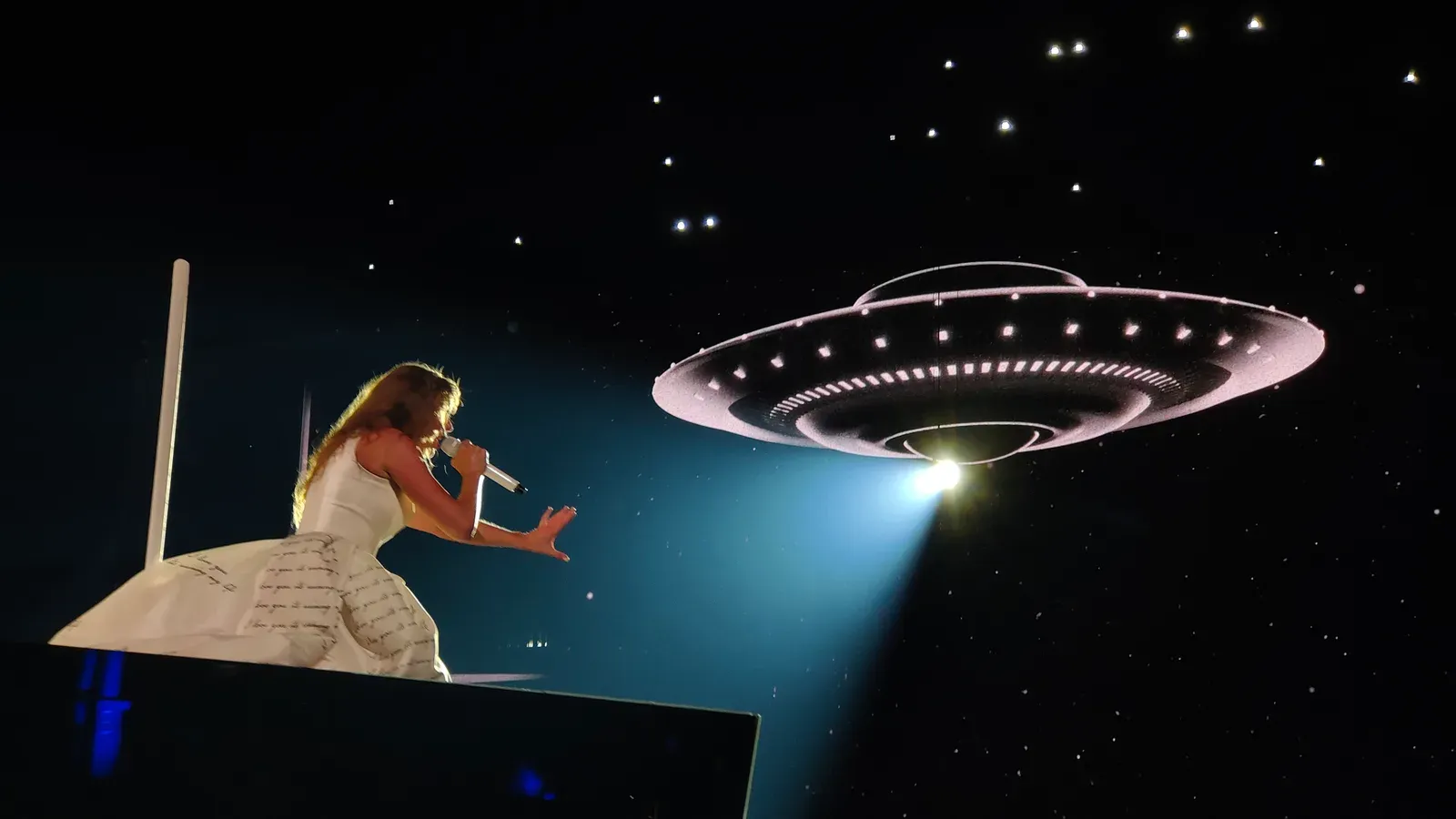 Taylor Swift ในชุดสีขาวร้องเพลงบนเวทีช่วงอัลบั้ม The Tortured Poets Department ของคอนเสิร์ต The Eras Tour ที่ Paris La Défense Arena 11 พฤษภาคม 2024 โดยมีภาพยานอวกาศบนจอด้านหลัง