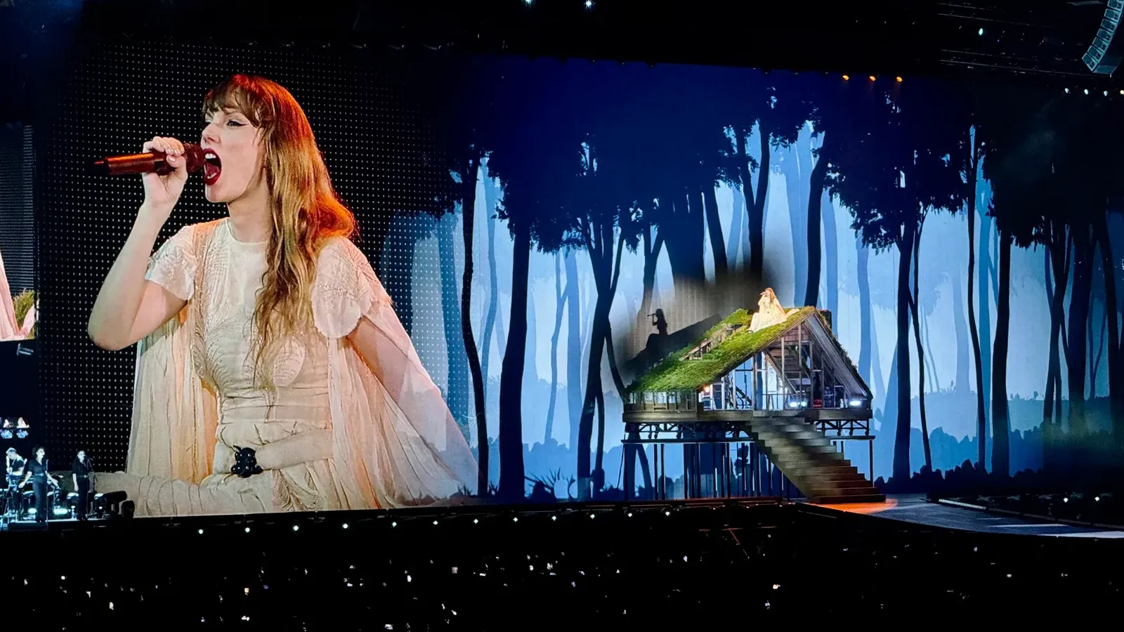 Taylor Swift บนจอยักษ์ขณะร้องเพลงในคอนเสิร์ต The Eras Tour ที่ Singapore National Stadium 9 มีนาคม 2024 ข้างฉากบ้านหลังคามอสกลางป่า