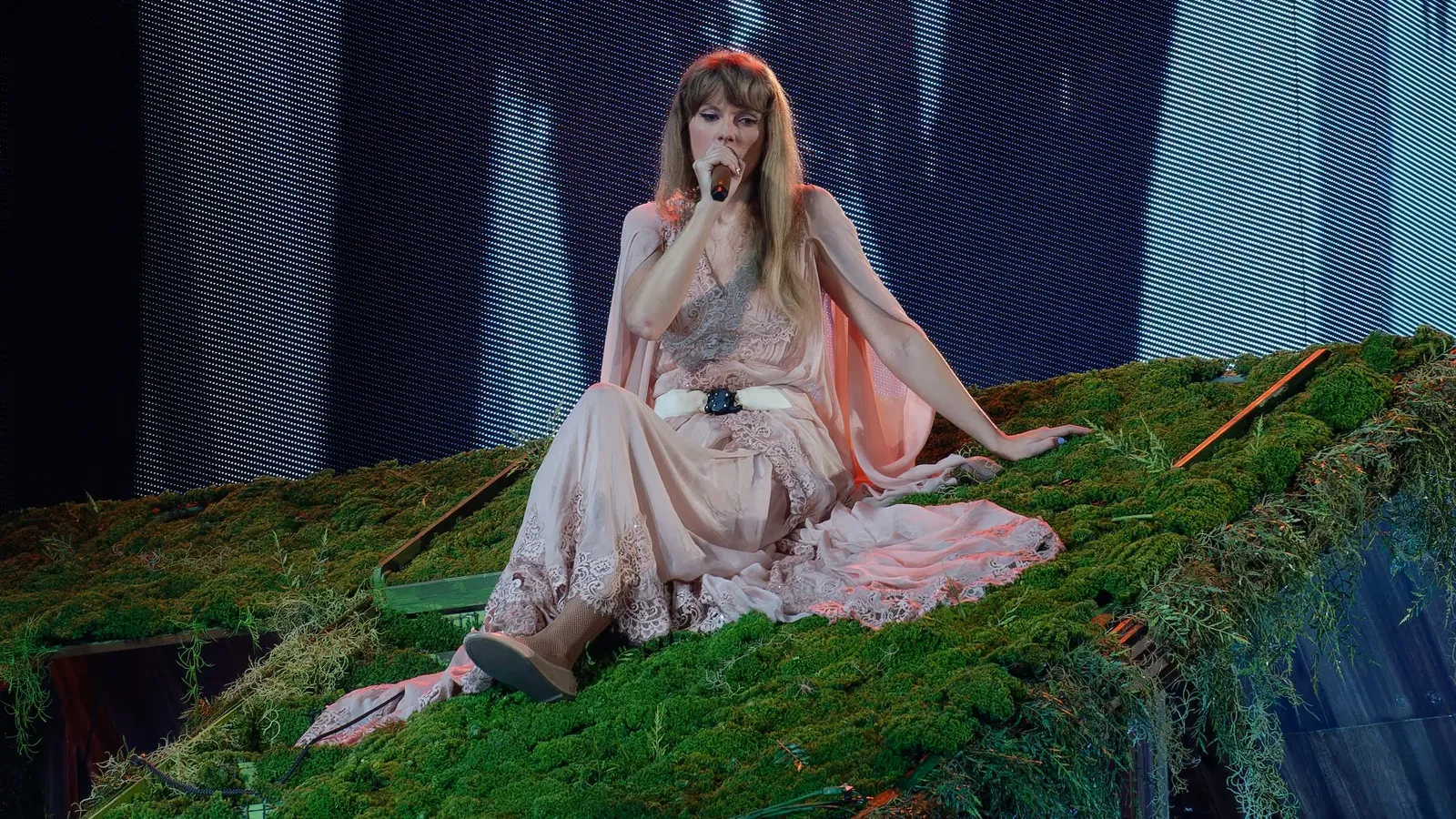 Taylor Swift ในชุดเดรสสีชมพูนั่งบนเวทีที่ปูด้วยมอสสีเขียวและร้องเพลงในคอนเสิร์ต The Eras Tour ที่เมือง Arlington รัฐเท็กซัส 2 เมษายน 2023