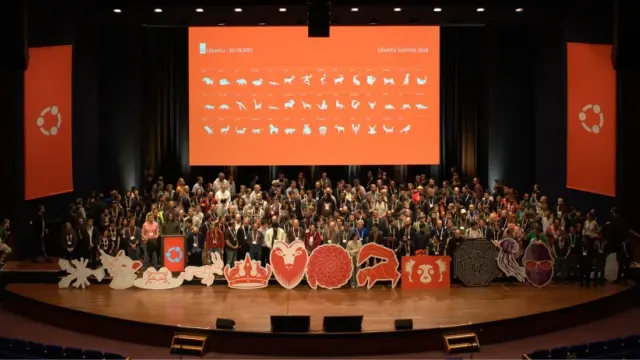 ผู้คนจำนวนมากรวมตัวบนเวทีที่มีธงและจอสีส้มของ Ubuntu