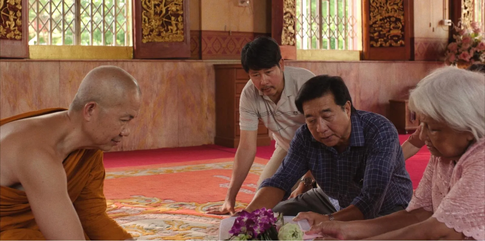 ภาพนิ่งจากภาพยนตร์ 9 วัดสู่สวรรค์ รางวัล Sutherland Award ปี 2026 แสดงคุณย่าและสมาชิกครอบครัวนั่งต่อหน้าพระภิกษุบนพรมในโบสถ์ มีดอกกล้วยไม้สีม่วงวางอยู่ด้านหน้า