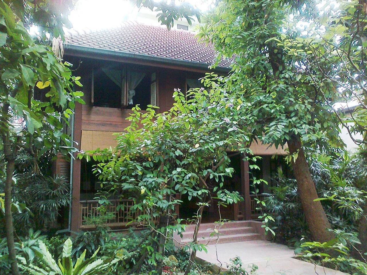 เรือนไม้สองชั้นสีน้ำตาลในพิพิธภัณฑ์ชาวบางกอก ซ่อนอยู่หลังต้นไม้