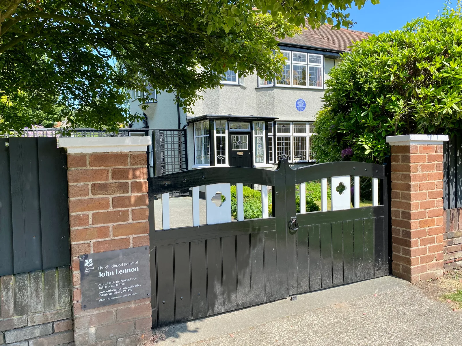 ประตูรั้วหน้า Mendips กับป้ายของ National Trust ที่เขียนว่า The childhood home of John Lennon เห็นสวนหน้าบ้านและระเบียงกระจกหลังรั้ว