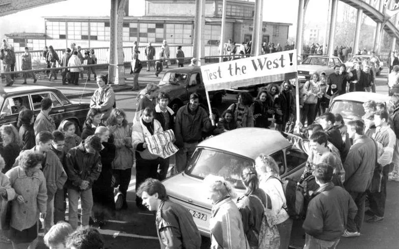 ฝูงชนยืนสองข้างถนนบนสะพานเหล็กที่ด่าน Bornholmer Straße ขณะรถยนต์ Trabant และรถคันอื่นแล่นผ่าน มีป้ายผ้าเขียนว่า Test the West! วันที่ 10 พฤศจิกายน 1989 หลังกำแพงเบอร์ลินเปิด