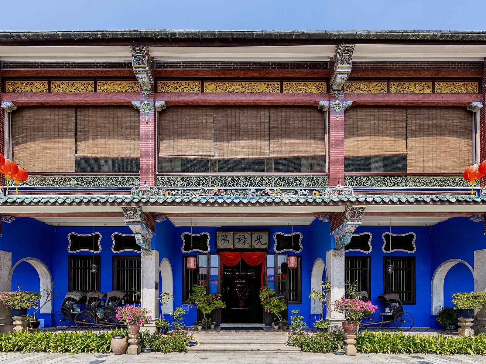 ด้านหน้า Blue Mansion ปีนัง หรือคฤหาสน์ Cheong Fatt Tze ผนังสีคราม ระเบียงชั้นสองมีราวเหล็กหล่อ และไม้กระถางเรียงหน้าบ้าน ภาพถ่ายเดือนมกราคม 2020