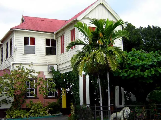 ตัวบ้านของ Bob Marley Museum ชั้นล่างก่ออิฐ ชั้นบนเป็นผนังไม้สีขาว หน้าต่างบานเกล็ดสีแดง หลังคาสังกะสีสีแดง และต้นปาล์มหน้าบ้าน