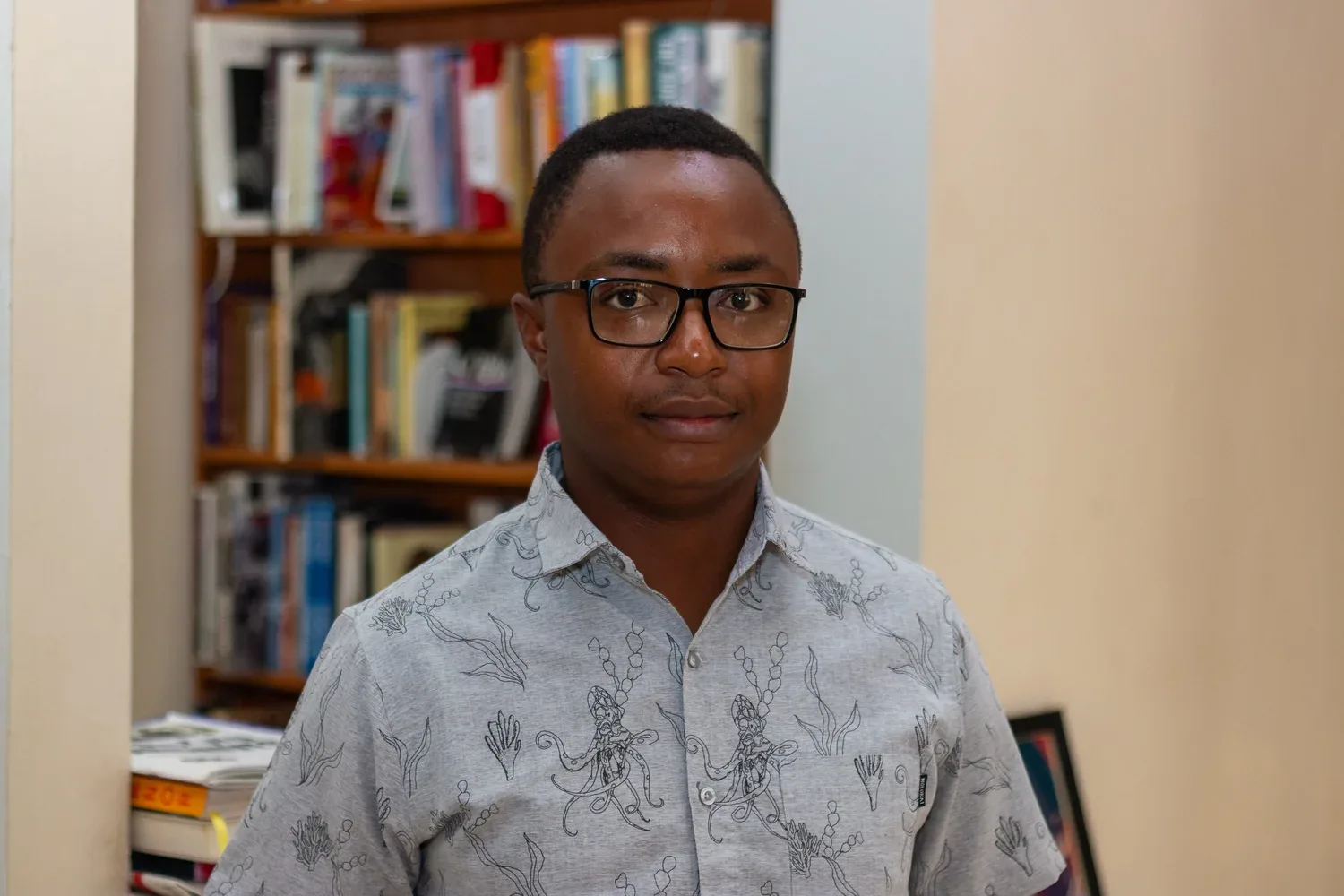 Simbarashe Steyn Kundizeza ผู้ชนะ Caine Prize 2026 สวมแว่นและเสื้อเชิ้ตลายพิมพ์ ยืนหน้าชั้นหนังสือ
