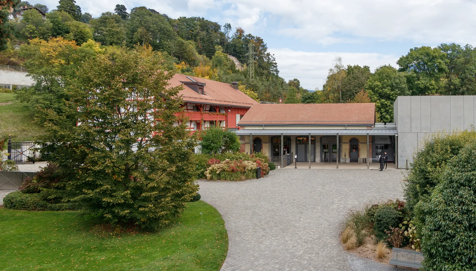 ลานทางเข้าพิพิธภัณฑ์ Chaplin's World ในที่ดิน Manoir de Ban อาคารหลังคากระเบื้องสีส้ม อาคารคอนกรีตทางขวา และสวนไม้พุ่มรอบลาน