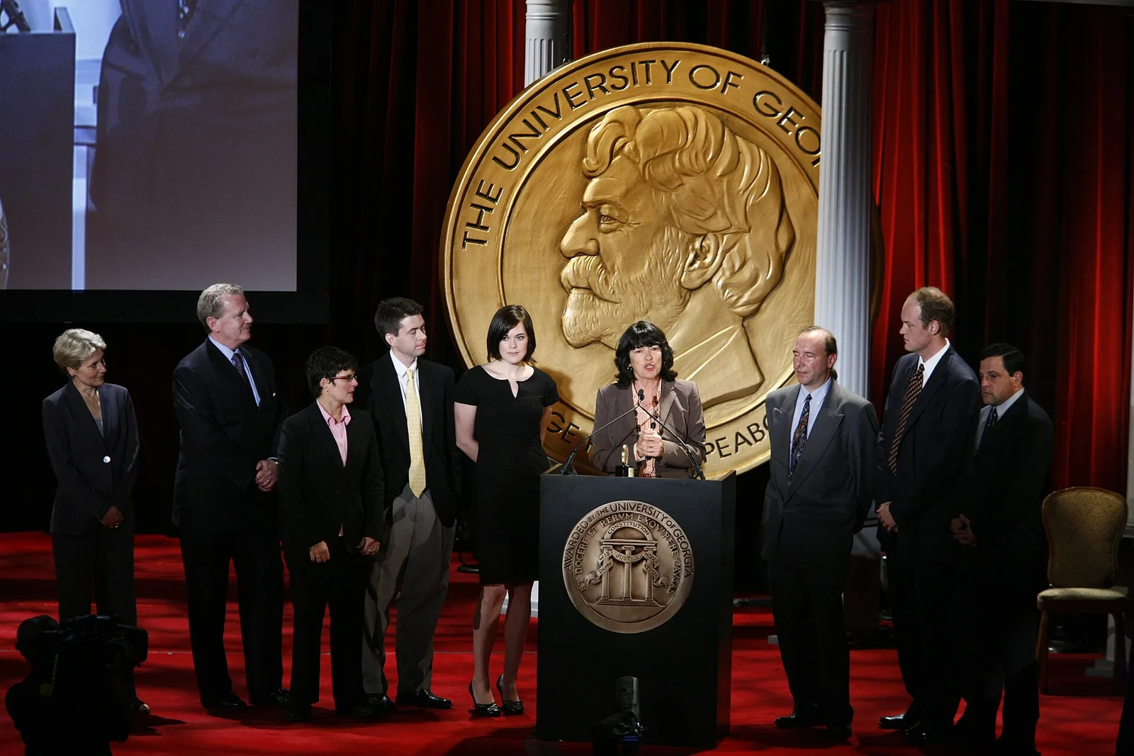Christiane Amanpour พูดที่โพเดียมหน้าเหรียญรางวัล Peabody ขนาดใหญ่ โดยมีทีมงานยืนเรียงแถว ในงาน Peabody Awards ครั้งที่ 67 ปี 2008