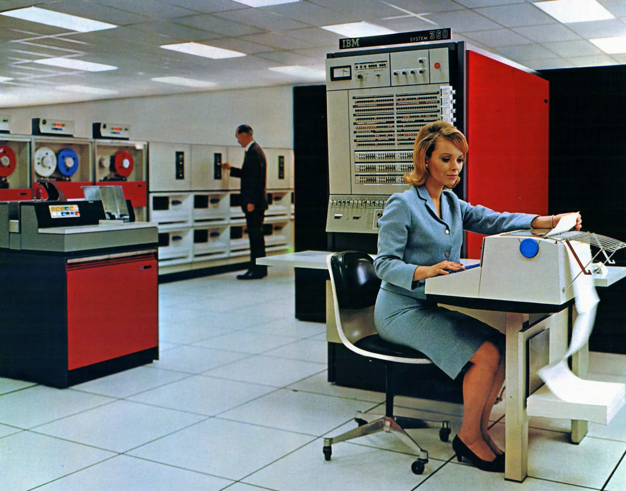 ผู้หญิงนั่งที่เครื่องพิมพ์ของเมนเฟรม IBM System/360 Model 65 หน้าตู้หน่วยประมวลผลกลางที่มีแผงควบคุมอยู่ด้านหน้า