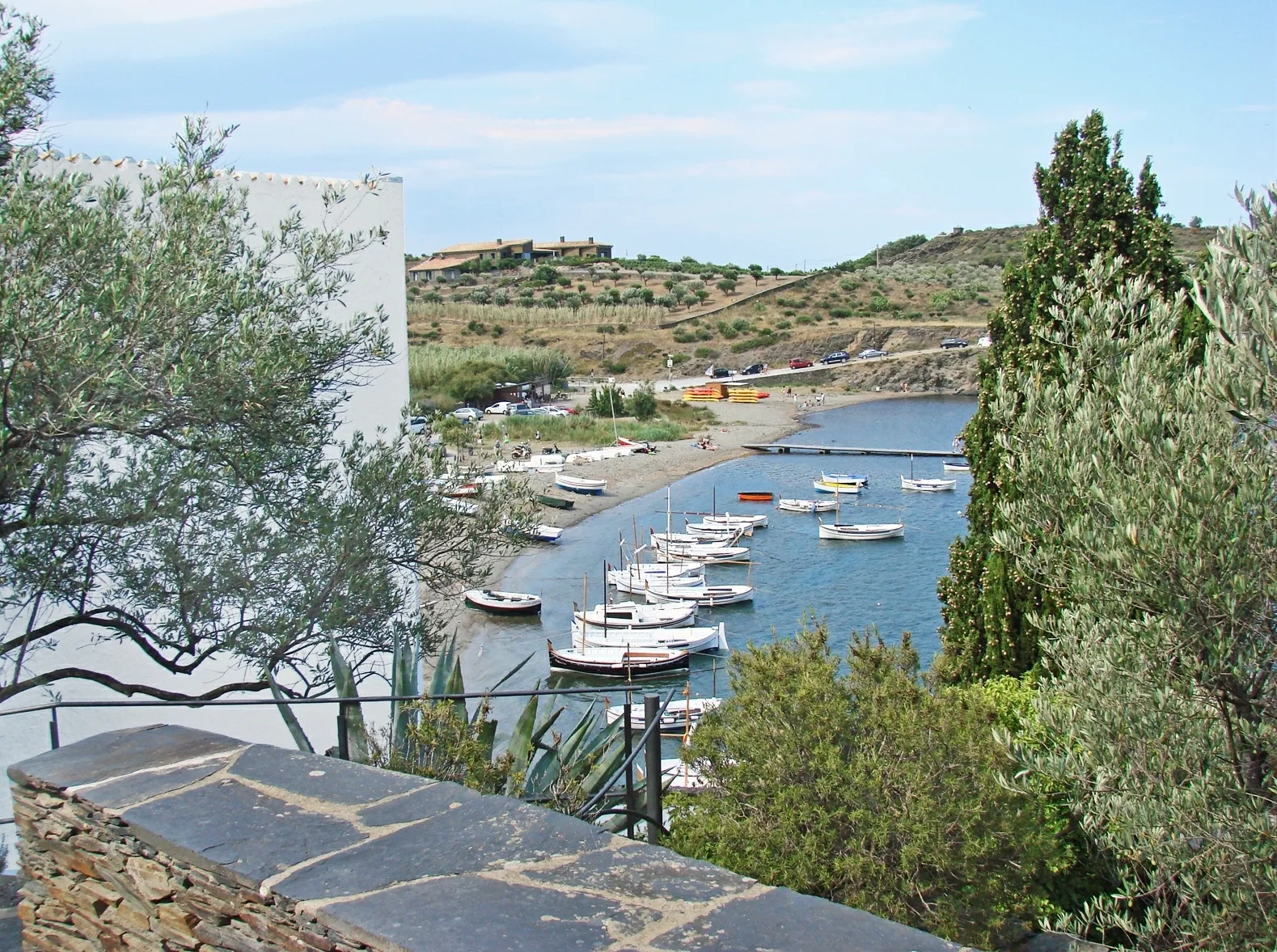 อ่าว Portlligat มองจากลานของบ้านพิพิธภัณฑ์ Salvador Dalí เห็นเรือเล็กจอดในอ่าว เนินเขาฝั่งตรงข้าม และผนังสีขาวของบ้านทางซ้าย ภาพถ่ายเดือนกรกฎาคม 2014
