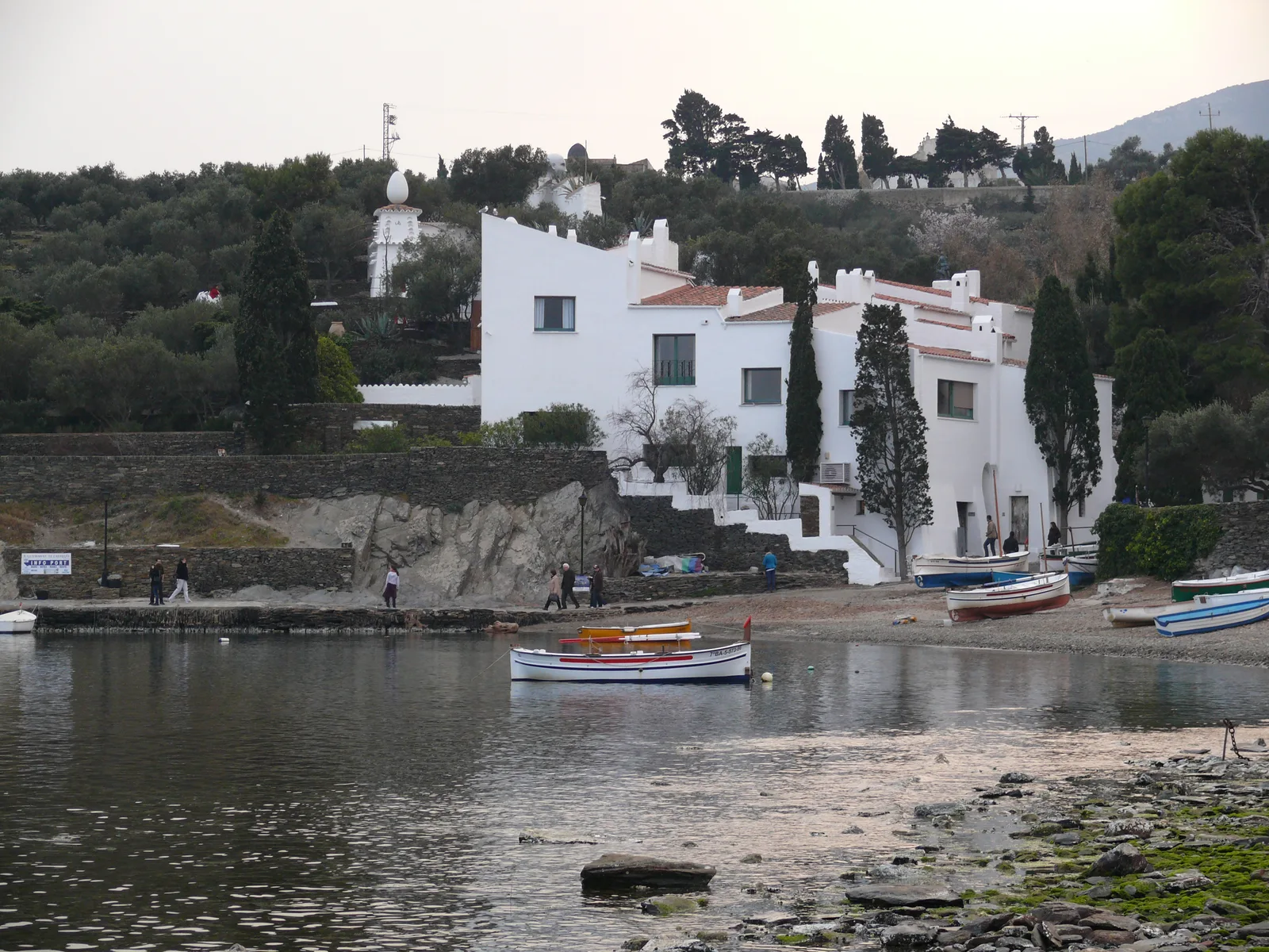 Casa Salvador Dalí ที่ Portlligat มองจากชายหาด อาคารสีขาวหลายระดับไล่ขึ้นเนิน มีต้นไม้ทรงสูงและเรือเล็กจอดหน้าบ้าน ภาพถ่ายเดือนมีนาคม 2012