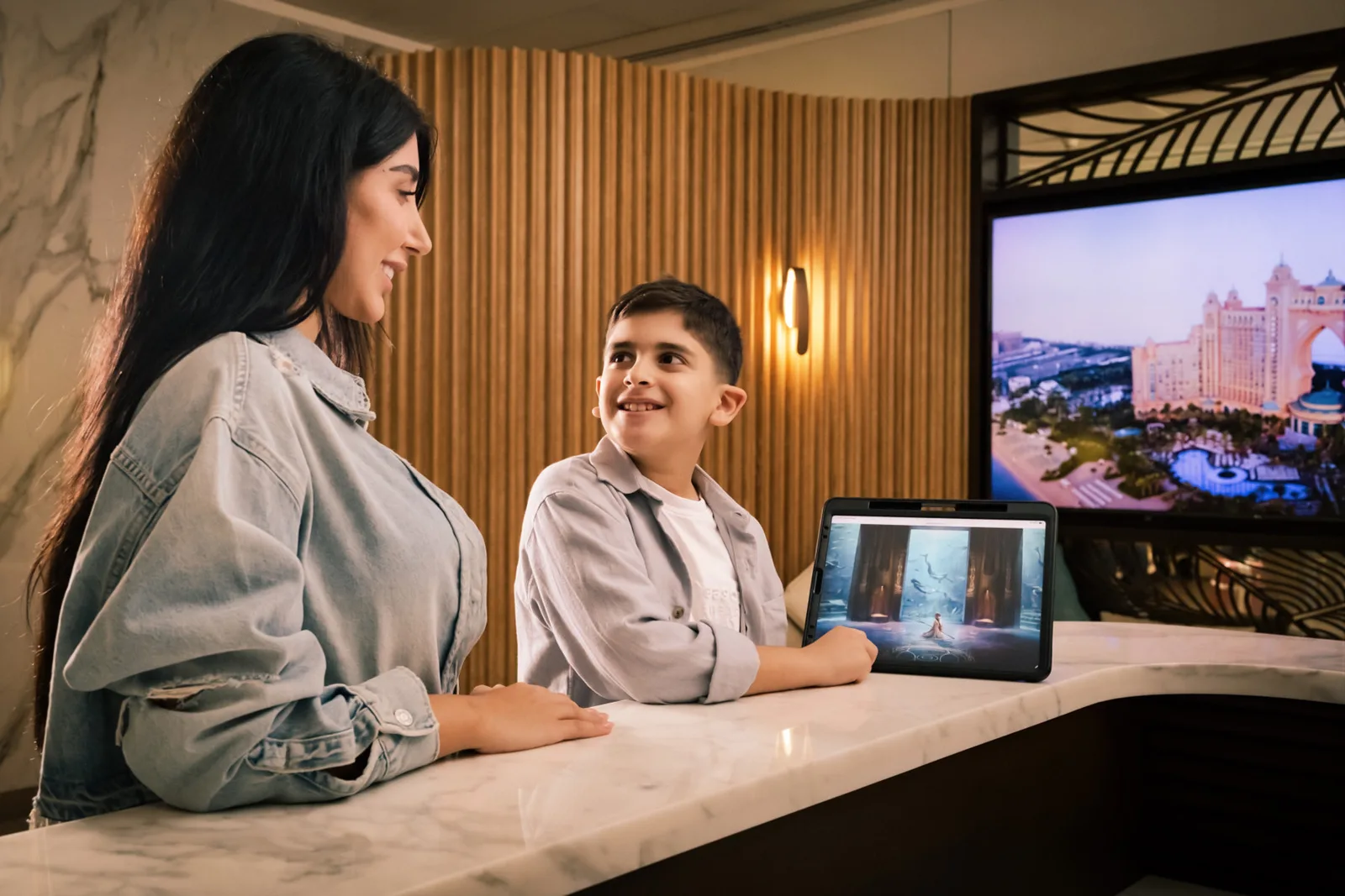 ภาพประกอบประกาศ Emirates Mile-A-Minute 2026 ของ Emirates แสดงแขกที่เคาน์เตอร์ต้อนรับ มีภาพโรงแรม Atlantis The Palm ในดูไบบนจอด้านหลัง