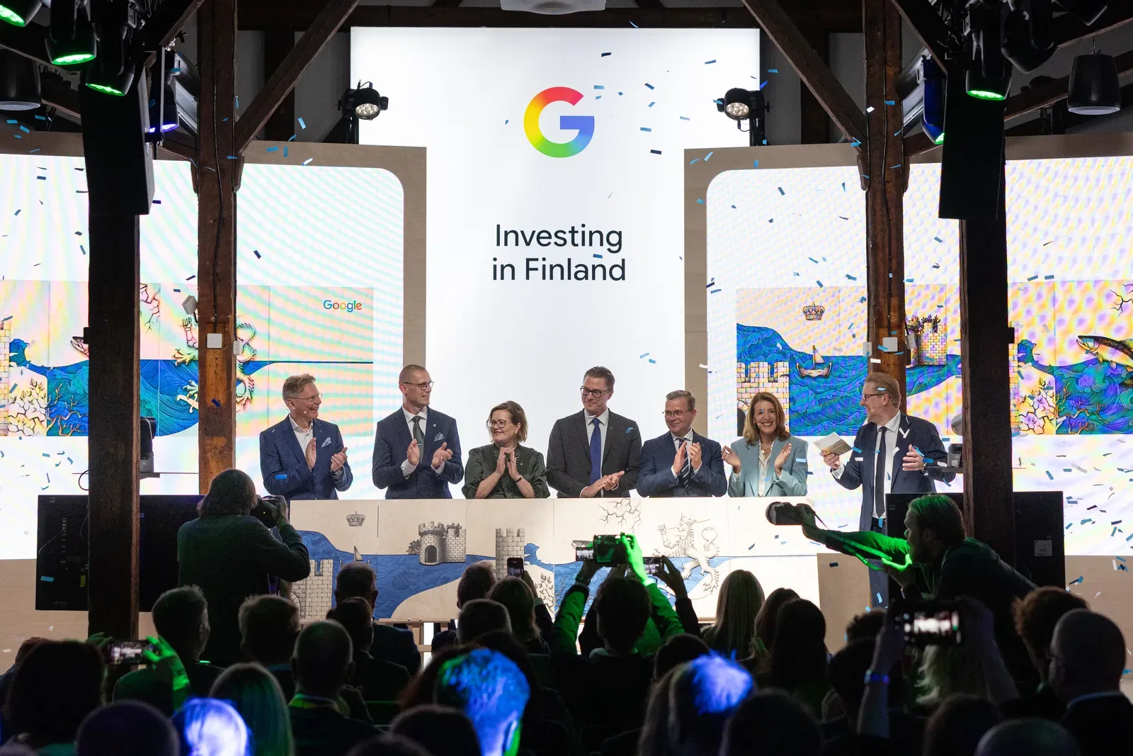 ผู้ร่วมงานเจ็ดคนบนเวทีหน้าจอข้อความ Investing in Finland ในงานประกาศลงทุน 13,000 ล้านยูโรของ Google ในฟินแลนด์