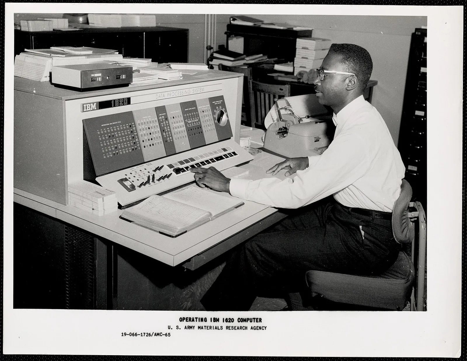 เจ้าหน้าที่กดสวิตช์บนคอนโซลของ IBM 1620 ซึ่งมีแผงไฟแสดงสถานะ ข้างเครื่องพิมพ์ดีดของระบบ ในภาพถ่ายของกองทัพบกสหรัฐฯ ราวปี 1963–1967