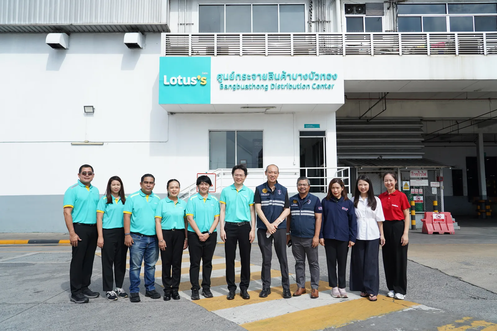 คณะเจ้าหน้าที่ยืนหน้าอาคารที่มีป้าย Lotus's ศูนย์กระจายสินค้าบางบัวทอง (Bangbuathong Distribution Center)
