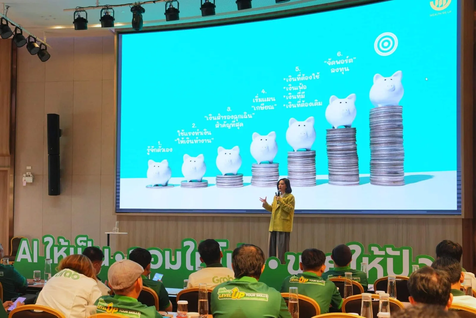 ห้องอบรม GrabAcademy คนขับในเสื้อ Grab นั่งฟังวิทยากรบนเวที หน้าจอสไลด์ภาษาไทย 6 ขั้นด้านการเงิน ภาพกระปุกออมสินบนกองเหรียญ และโลโก้ Wealth Me Up