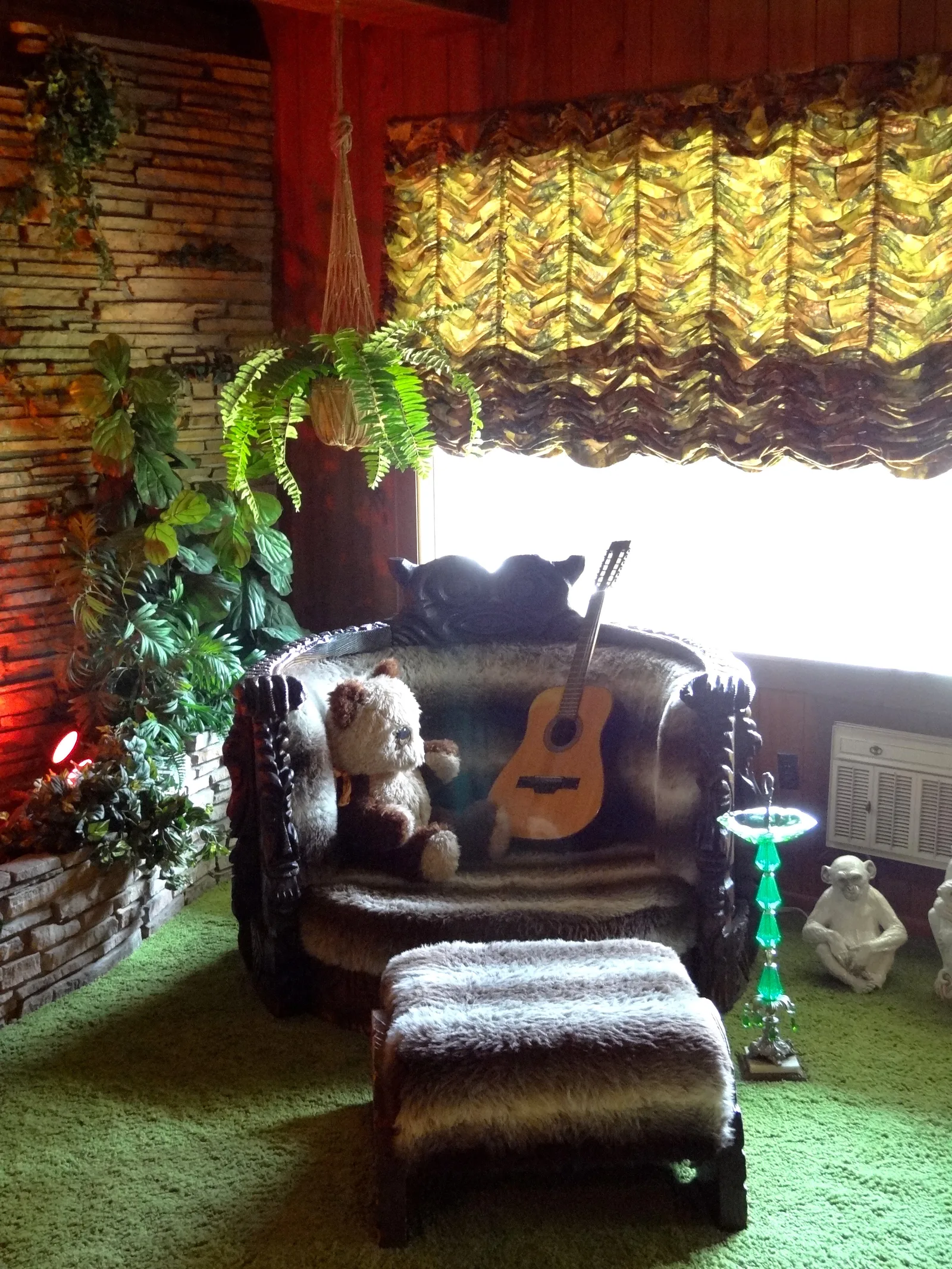 Jungle Room ในเกรซแลนด์ มีเก้าอี้ไม้แกะสลักบุขนสัตว์เทียม พรมขนยาวสีเขียว ต้นไม้แขวน และผนังหินทางซ้าย ภาพถ่ายเมื่อพฤษภาคม 2012