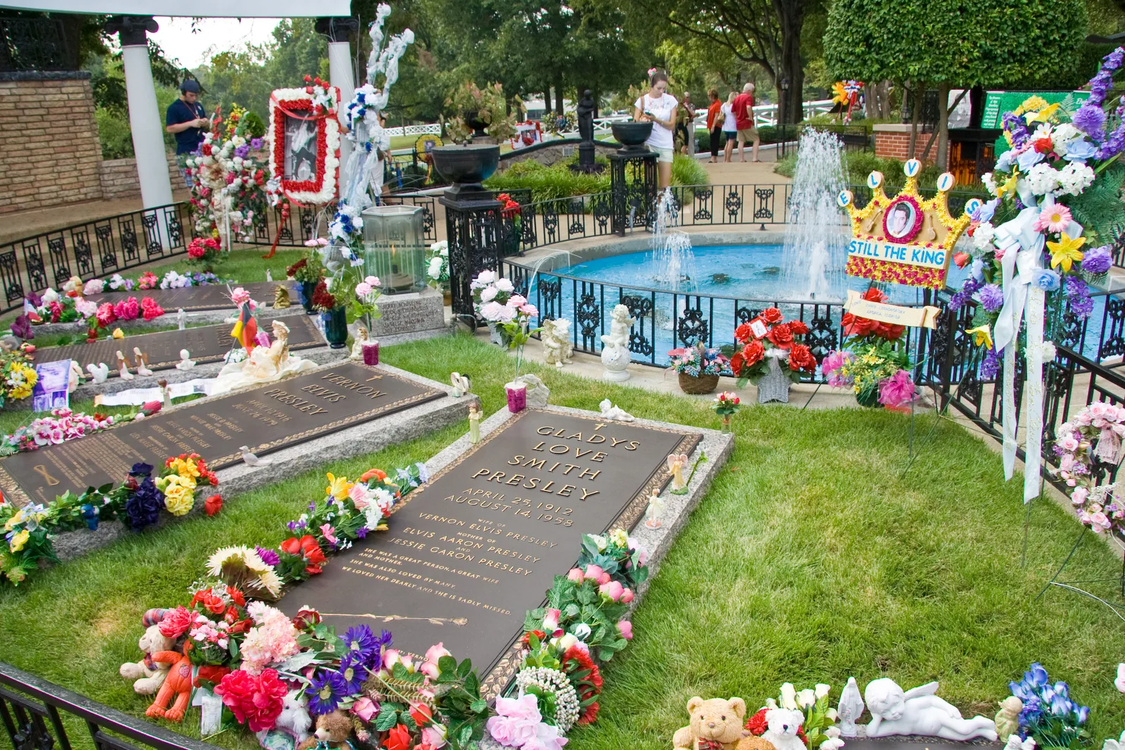 หลุมศพของครอบครัว Presley ใน Meditation Garden ของเกรซแลนด์ ประดับดอกไม้และของระลึกจากแฟนเพลง ด้านหลังเป็นสระน้ำพุล้อมรั้วเหล็ก