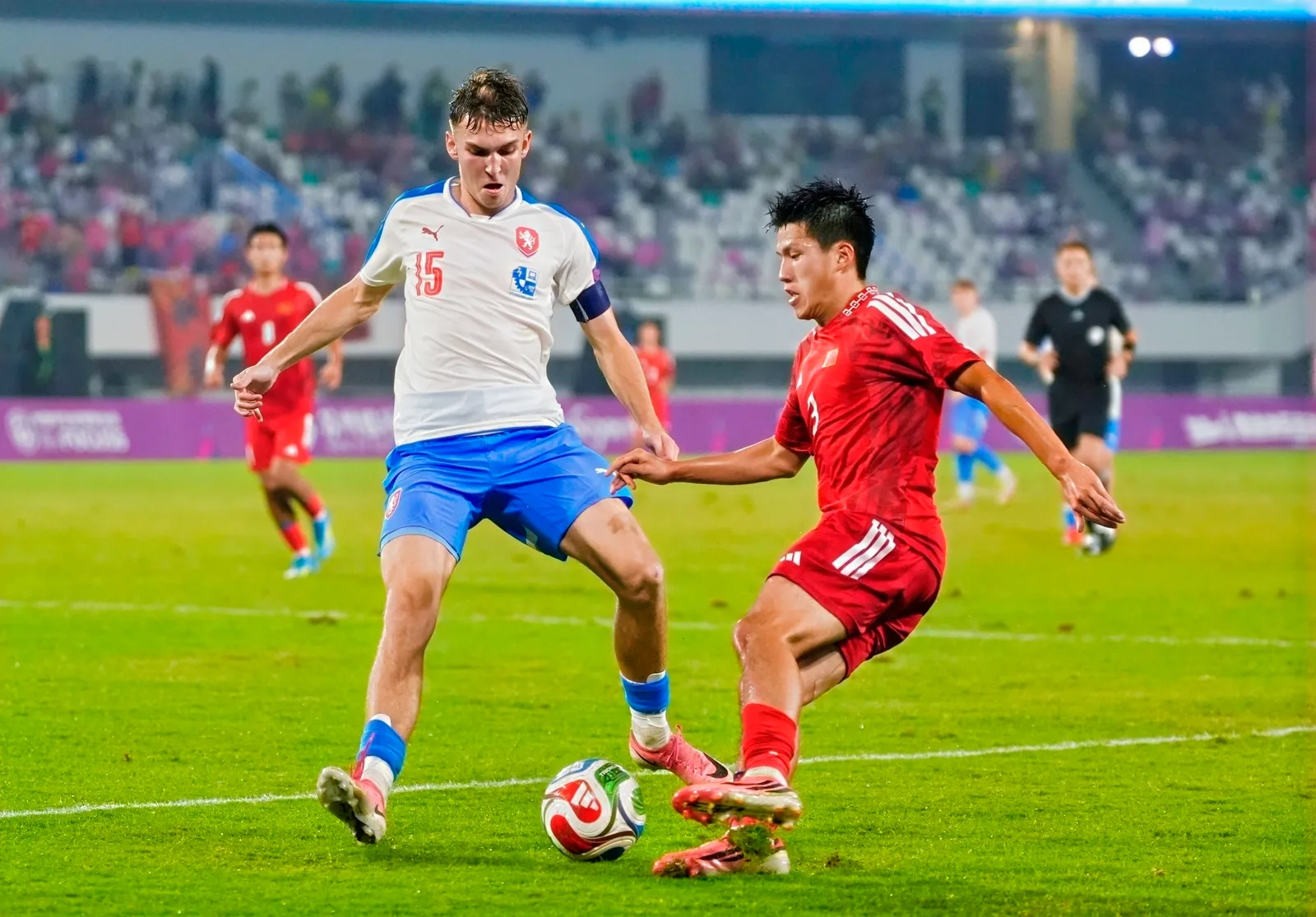 นัดเปิดสนามฟุตบอลนักเรียนชิงแชมป์โลก 2026: ผู้เล่น China 1 ในชุดแข่งสีแดงเลี้ยงบอลหนีผู้เล่น Czechia 2 ในชุดสีขาวกางเกงสีฟ้า