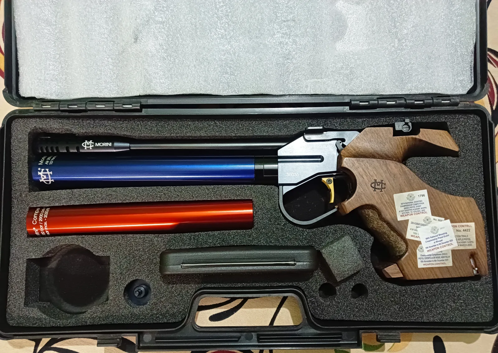 ปืนสั้นอัดลมกีฬา Morini 162 ด้ามไม้ ถังอากาศสีน้ำเงิน และถังอากาศสำรองสีส้ม วางในกล่องบุโฟม อุปกรณ์แบบที่ใช้ในประเภทปืนสั้นอัดลม 10 เมตร
