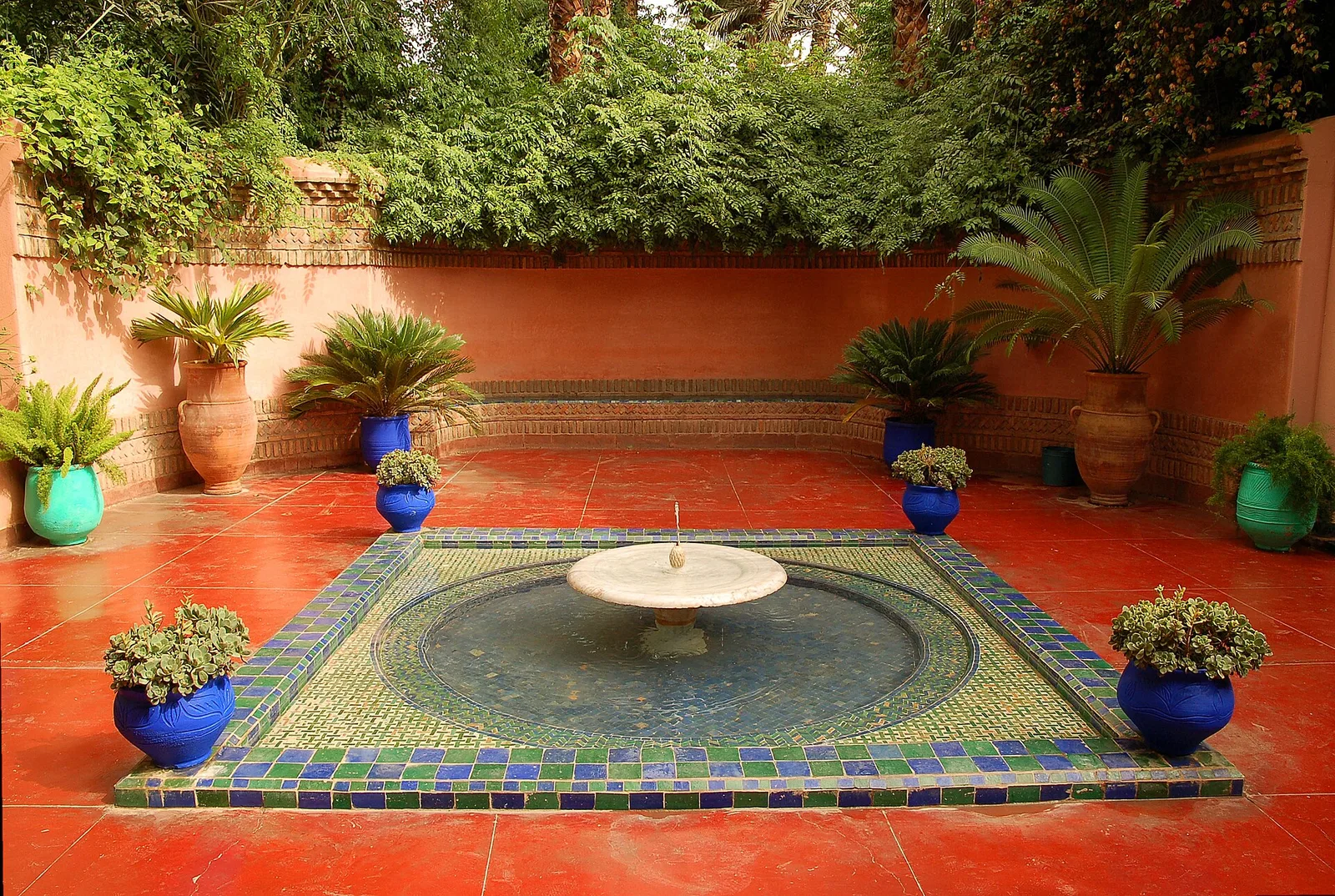 ลานน้ำพุทรงกลมปูกระเบื้องโมเสก ล้อมด้วยกระถางต้นไม้สีน้ำเงินใน Jardin Majorelle ภาพถ่ายเมื่อ 10 กรกฎาคม 2006