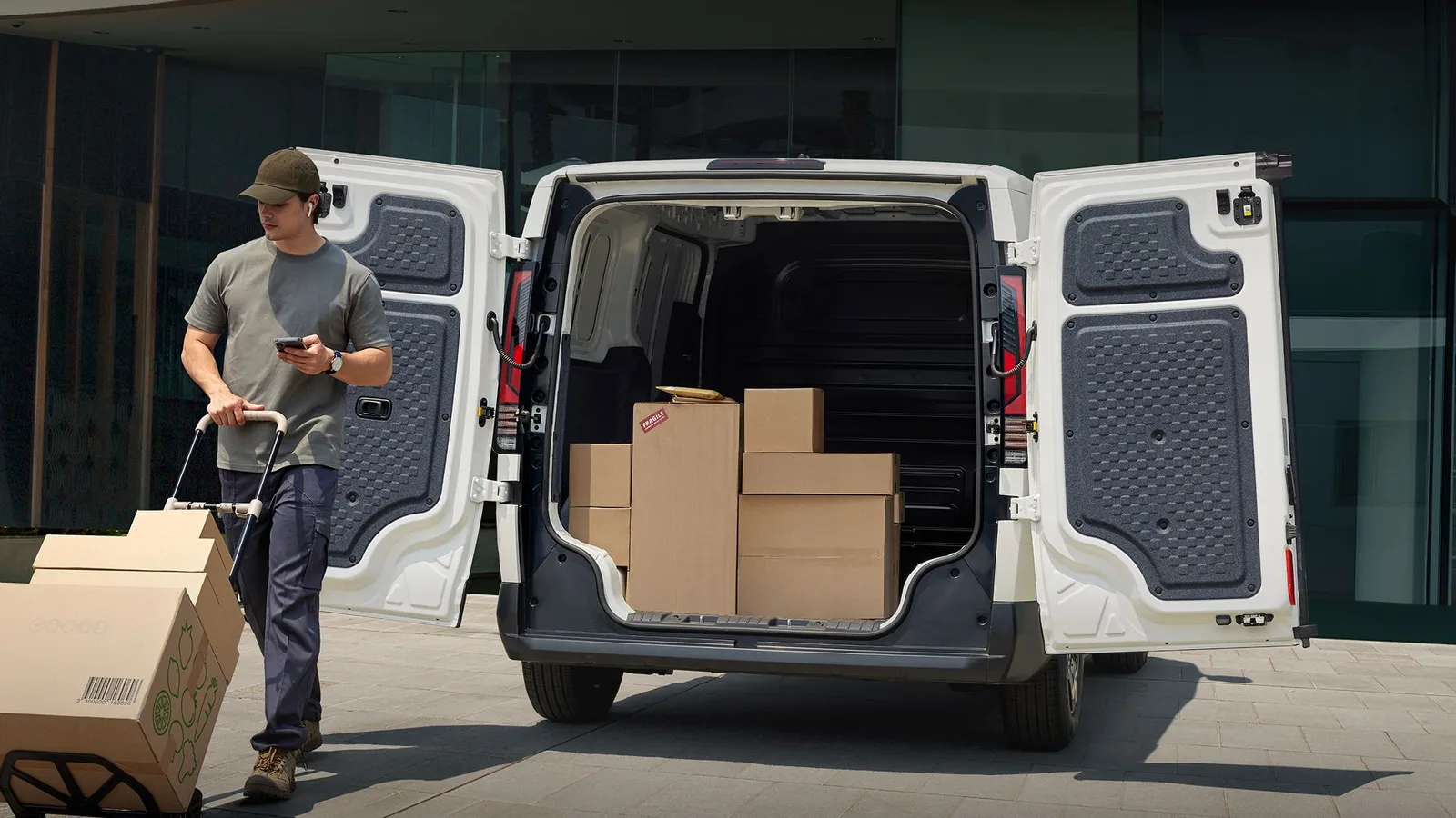 รถตู้ Kia PV5 Cargo สีขาวเปิดประตูหลัง มีกล่องสินค้าวางซ้อนในห้องบรรทุก และชายคนหนึ่งเข็นรถเข็นกล่องอยู่ข้างรถ