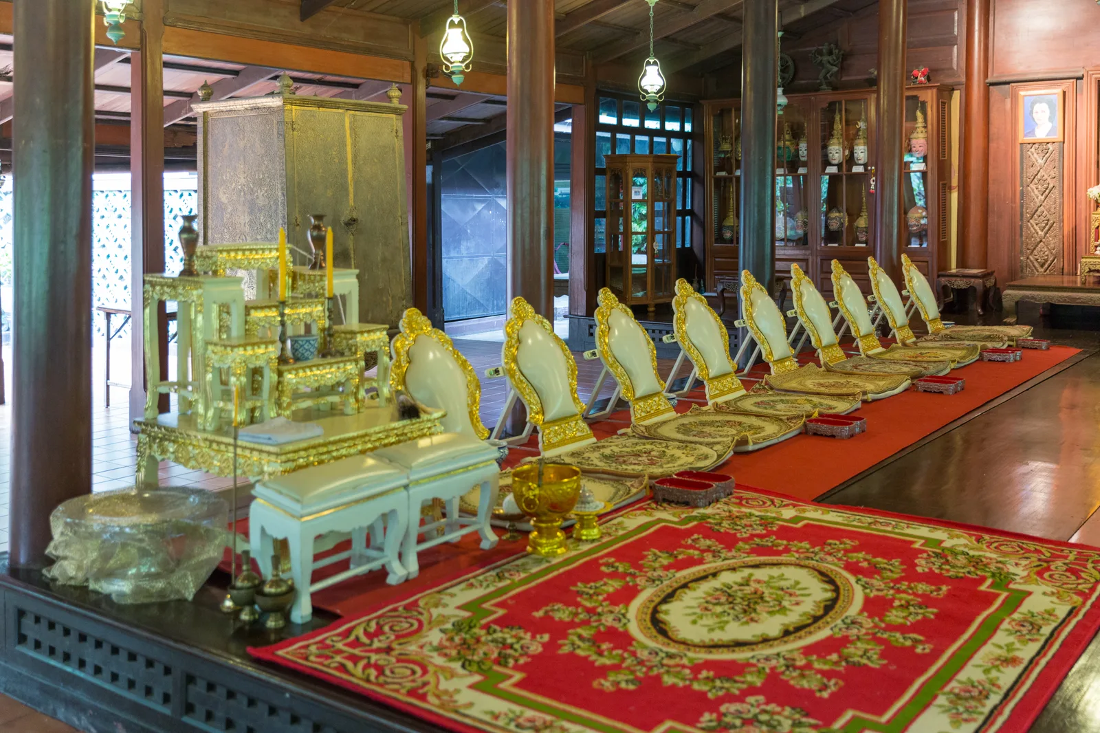 ห้องโถงในเรือนไม้ของบ้านคึกฤทธิ์ ซอยสวนพลู พื้นปูพรมแดง มีโต๊ะหมู่บูชาสีทองและเก้าอี้ปิดทองเรียงเป็นแถว ด้านหลังเป็นตู้ไม้จัดแสดงของ