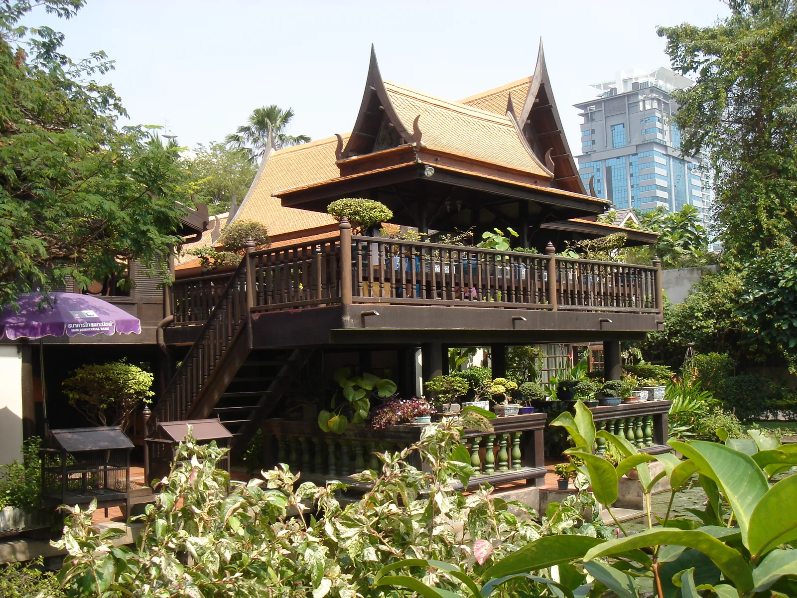 เรือนไทยไม้ยกพื้นสูงของบ้านคึกฤทธิ์ ซอยสวนพลู มีบันไดขึ้นชานชั้นบนที่ล้อมด้วยราวไม้และวางไม้กระถาง ด้านหลังเป็นอาคารสูง