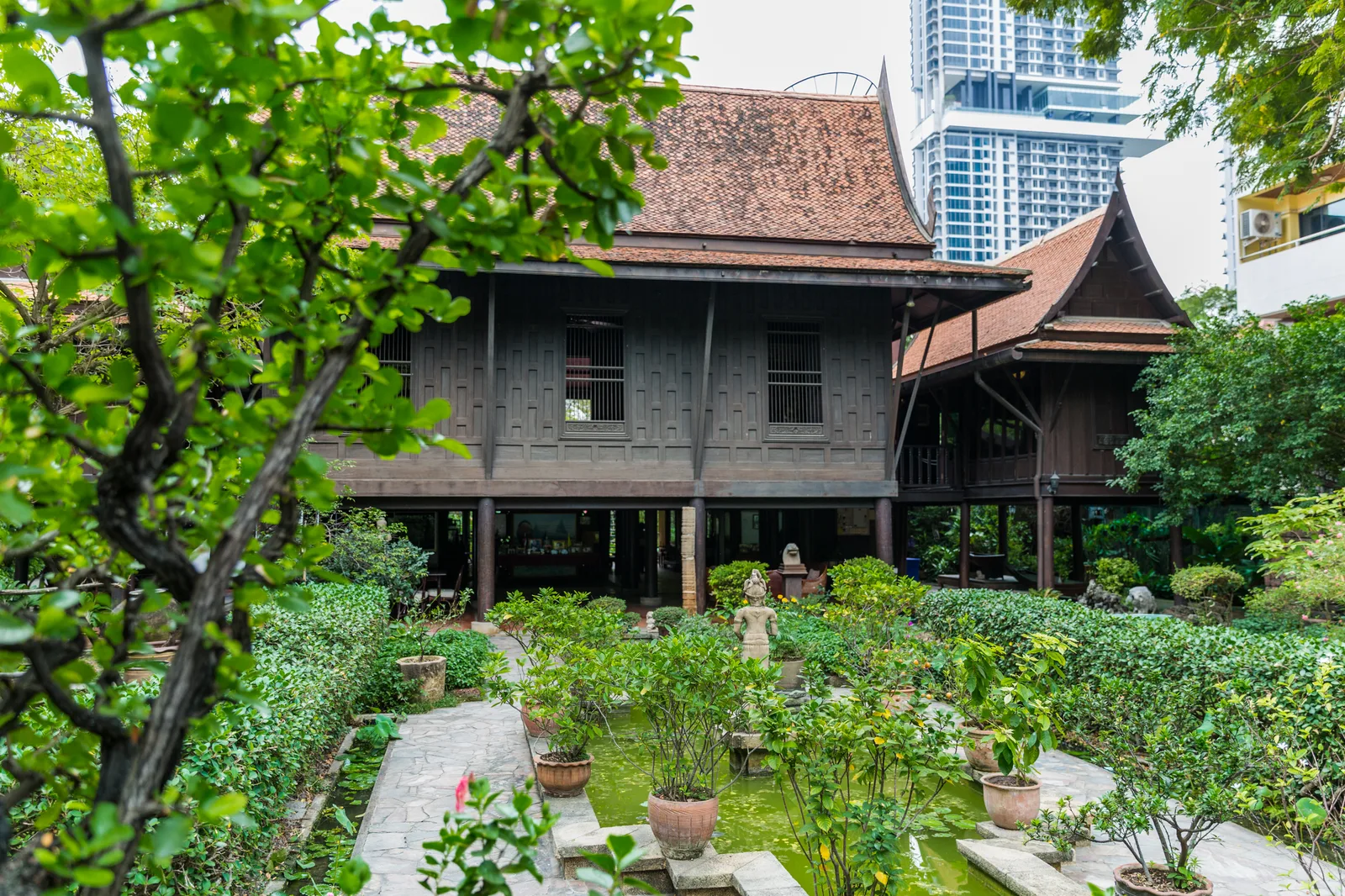 เรือนไทยไม้ยกพื้นสูงของบ้านซอยสวนพลู ฝาไม้สีเข้ม หลังคาทรงจั่วมุงกระเบื้อง หน้าเรือนเป็นทางเดินและไม้กระถาง มีอาคารสูงด้านหลัง