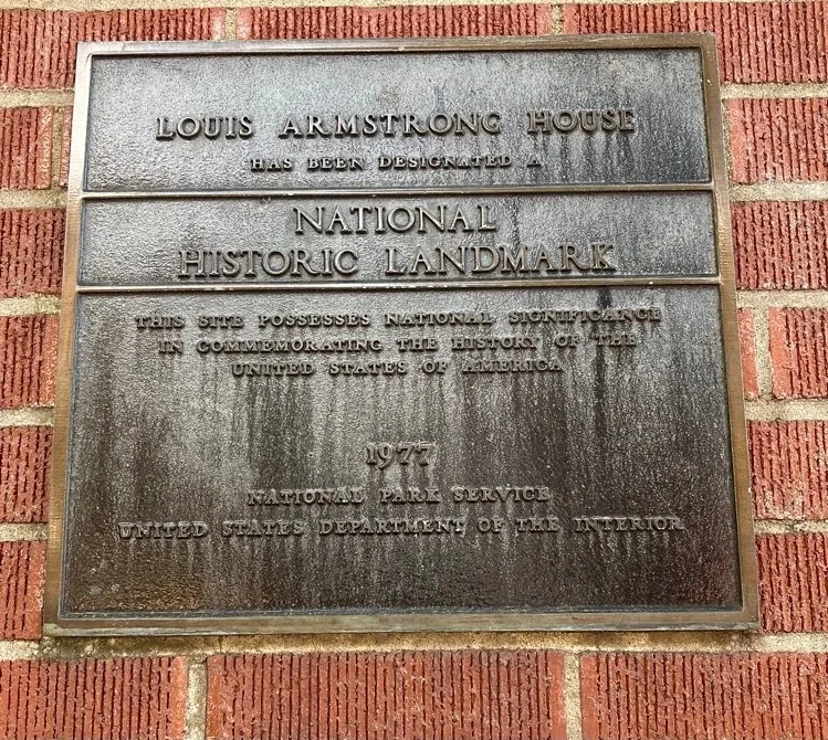 แผ่นป้ายโลหะบนผนังอิฐ ระบุว่า Louis Armstrong House ได้รับการประกาศเป็น National Historic Landmark ปี 1977 โดย National Park Service กระทรวงมหาดไทยสหรัฐฯ
