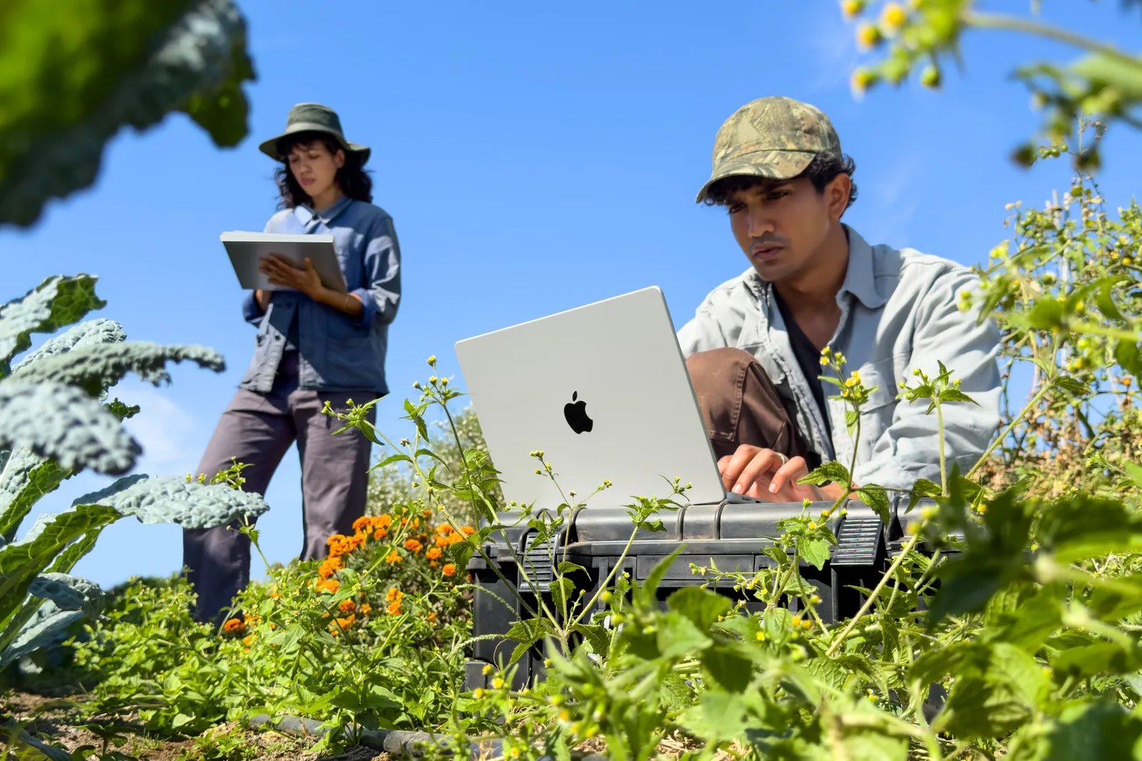 ผู้ใช้ MacBook Pro 14 นิ้ว ชิป M6 ทำงานกลางแปลงผักคู่กับเพื่อนร่วมงาน