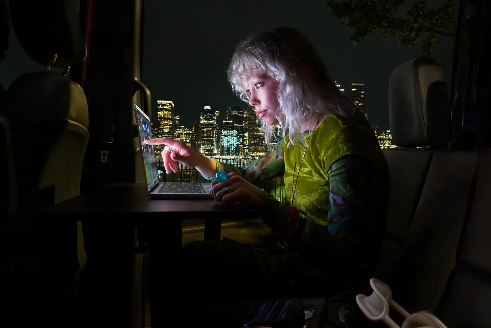 ผู้ใช้แตะหน้าจอ MacBook Pro จอ PROLED XDR ในที่มืด ด้านหลังเป็นทิวทัศน์เมืองยามค่ำ
